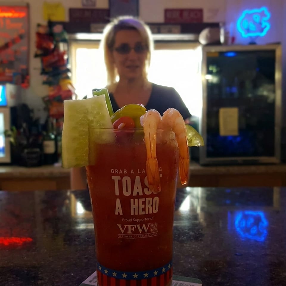 Red cocktail garnished with shrimp, citrus slices, and cucumber sits on a bar beside a bartender and glowing neon signs.