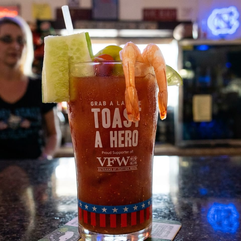 Bloody Mary garnished with shrimp, lime, cucumber, and celery sits on a bar, with a bartender and glowing signs in the background.
