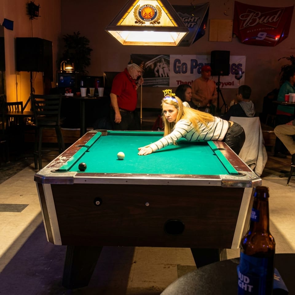 A woman plays pool in a warmly lit bar while patrons socialize and beer bottles sit in the foreground.