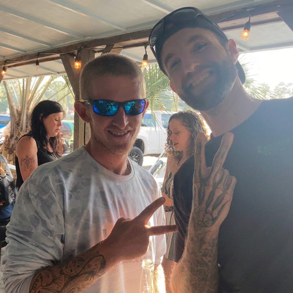Two tattooed men smile and pose on a sunlit restaurant patio, surrounded by other patrons and string lights.