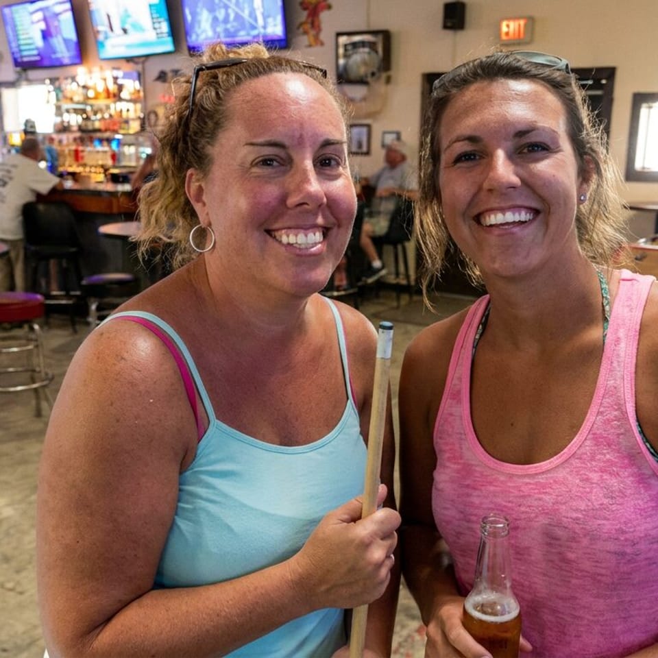 Two smiling women pose inside a lively bar, one holding a pool cue and the other holding a beer bottle, with foosball and TVs behind them.