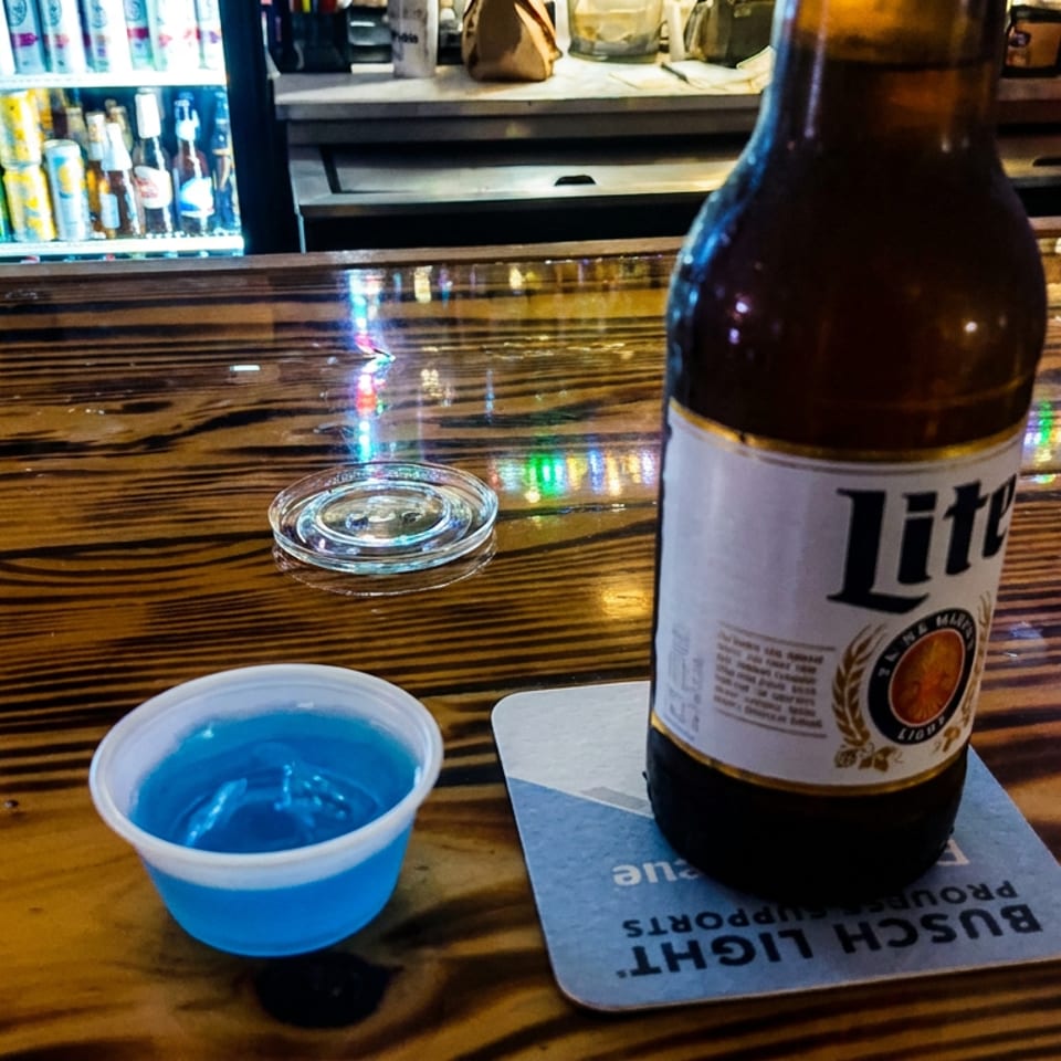 Lite beer bottle and blue drink in a plastic cup on a polished wooden bar beside an ashtray and beverage cooler.