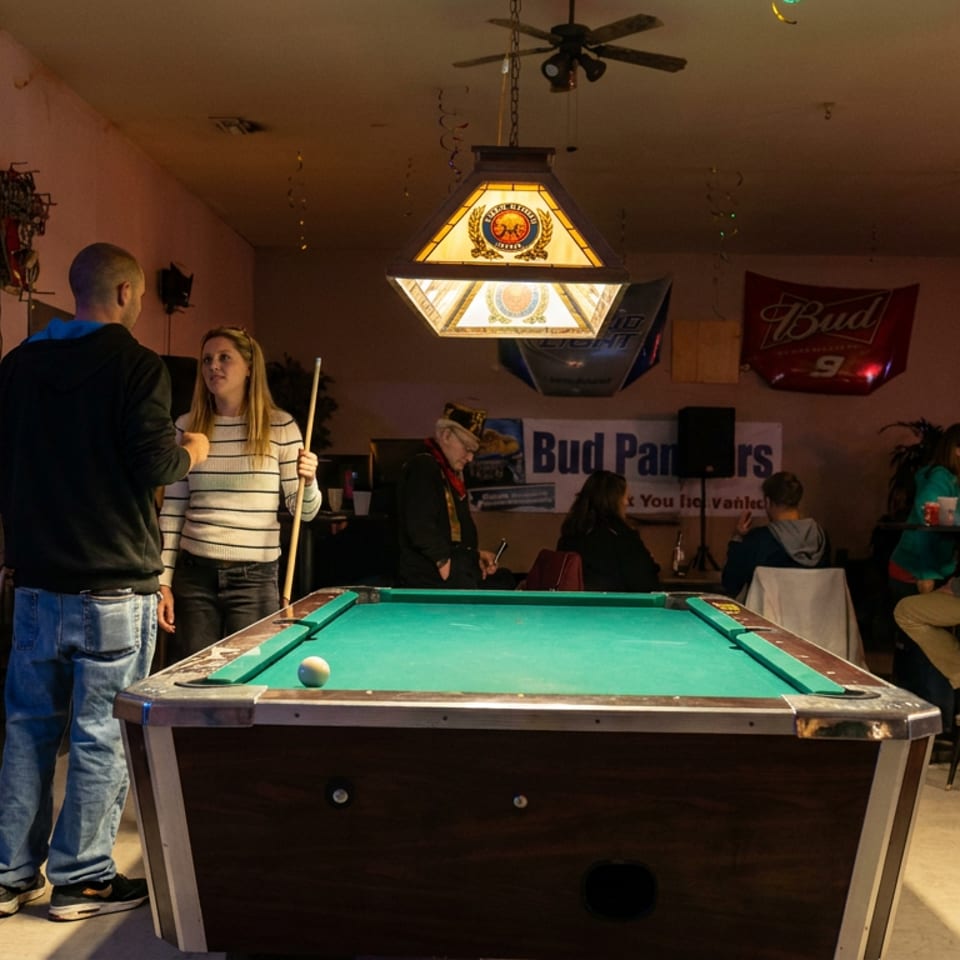 Warmly lit bar with patrons gathered around a green pool table, beer signs, ceiling fans, and a hanging billiards lamp.