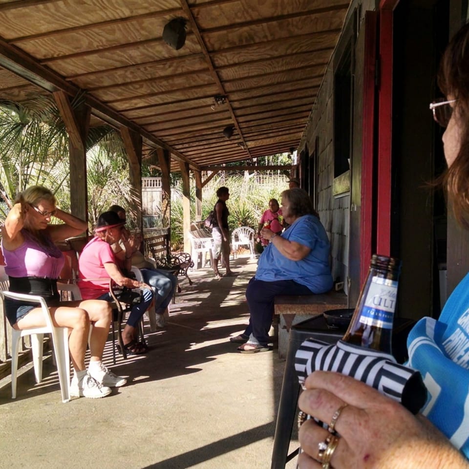 People socialize on a sunny, rustic covered patio, with palm trees, plastic chairs, and a woman holding a bottled drink.