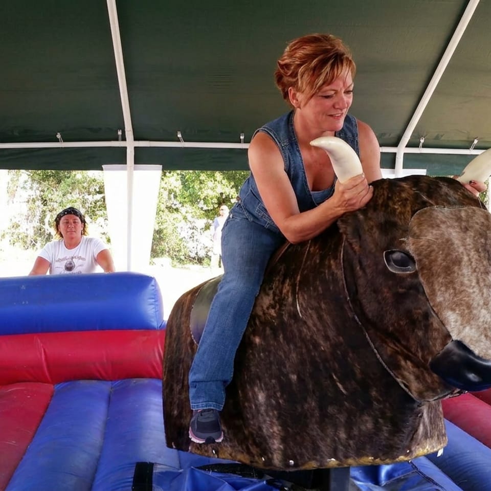Woman riding a mechanical bull inside a red and blue inflatable arena, with two people watching under a canopy.