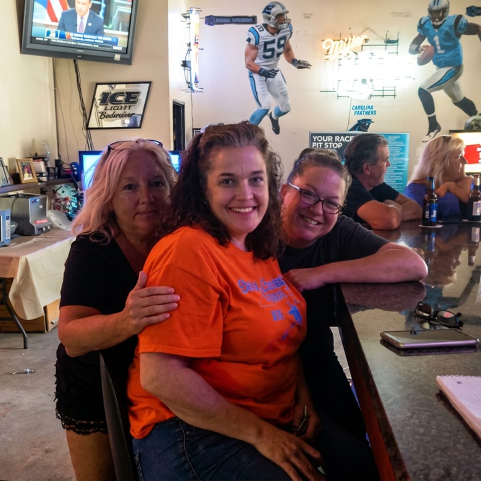 Three smiling people pose together inside a sports-themed restaurant with football murals, televisions, and patrons at the counter.
