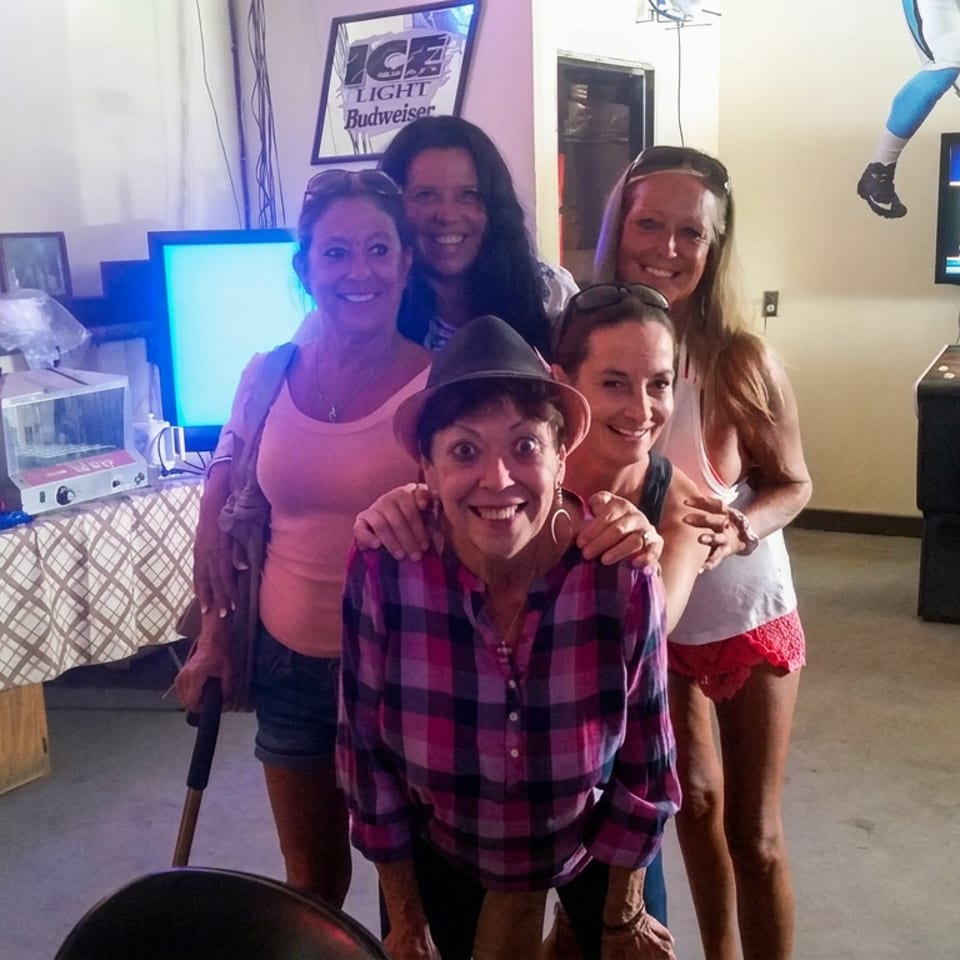 Five smiling women pose together inside a casual restaurant arcade beside games, memorabilia, and an American flag.