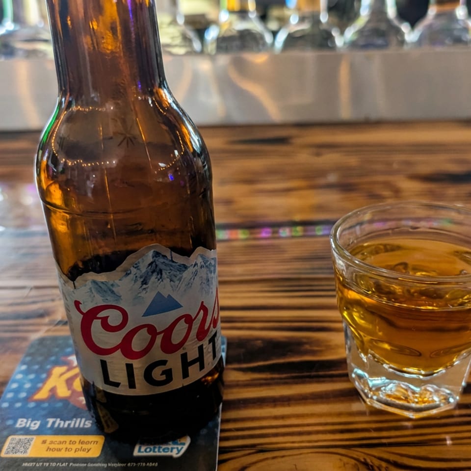 Coors Light beer bottle and whiskey glass on a wooden bar counter in a warmly lit restaurant.