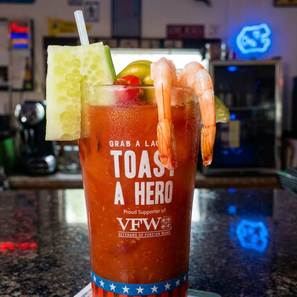 Bloody Mary cocktail garnished with shrimp, cucumber, tomato, lemon, and lime on a restaurant bar.